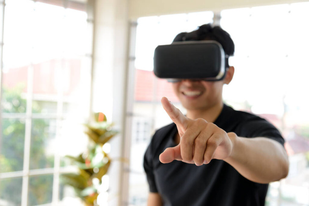 Nuori aasialainen mies saa kokemusta VR virtuaalitodellisuus kuulokkeet tai 3D-lasit sormella olohuoneessa kotona. tekniikka, pelaaminen, viihde käsite - Valokuva, kuva