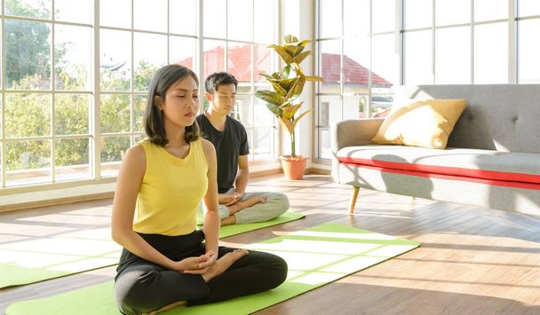Il est temps de pratiquer le yoga. Jeune couple asiatique assis à Ardha Padmasana exercice, les yeux fermés, portant des vêtements de sport sur le tapis à la maison. concept mode de vie - Photo, image