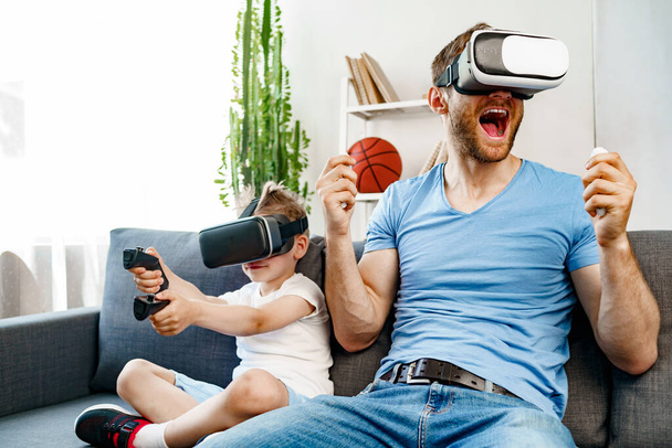 Dad and son sitting on sofa and playing games with VR glasses - Zdjęcie, obraz