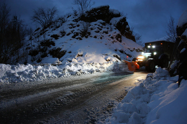 sneeuwschuiver passeert met lichten aan door de besneeuwde bergstraten in de avond om het wegverkeer op te ruimen - Foto, afbeelding