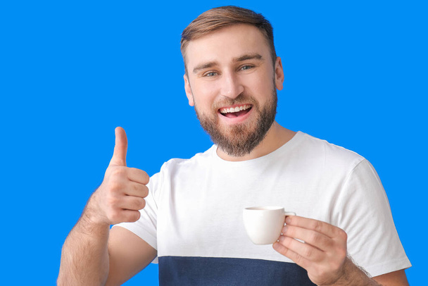 Przystojny młody człowiek z filiżanką gorącej kawy pokazuje kciuk do góry na tle koloru - Zdjęcie, obraz