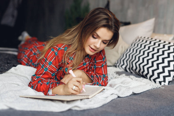 Blisko dziewczyny nosi uroczyste kłamstwa piżamy i pisać prawa ręka Lista życzeń w domu. Szczęśliwa kobieta w czerwonej koszuli nocnej na łóżku - Zdjęcie, obraz
