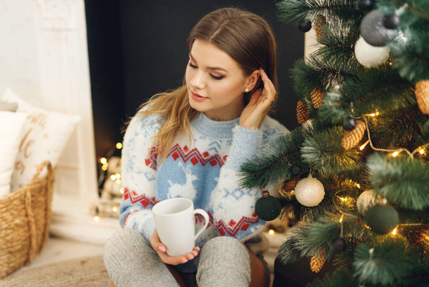 Il primo piano della ragazza indossa vestiti festivi prendere una tazza di caffè a casa vicino all'albero di Natale. Donna da sogno seduta su tappetino in maglione blu con cervo e calze grigie - Foto, immagini
