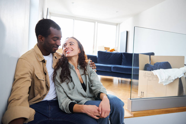 Young mixed ethnicity couple celebrating moving day in lounge of new home sitting on floor surrounded by removal boxes together - Photo, Image