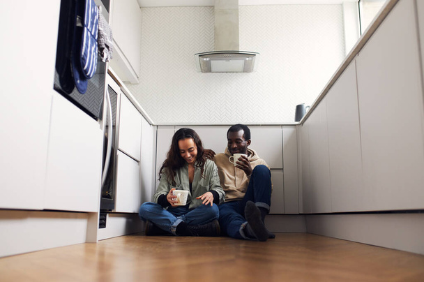 Young mixed ethnicity couple celebrating moving day in kitchen sitting on floor drinking hot drink together - Zdjęcie, obraz