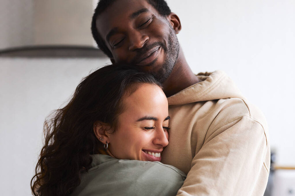 Loving young mixed ethnicity couple cuddling whilst standing in kitchen together - Foto, Imagem