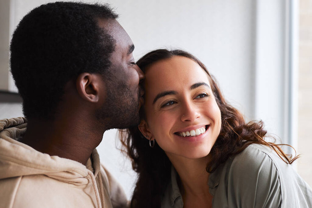 Loving young mixed ethnicity couple cuddling and kissing whilst standing in kitchen together - Photo, Image
