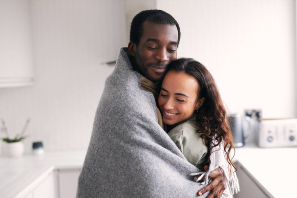Amar a la joven pareja étnica mixta abrazándose mientras están de pie en la cocina envueltos en una acogedora manta juntos - Foto, Imagen
