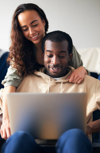 Γυναίκα αγκαλιάζει τον άνδρα από πίσω ως νεαρό ζευγάρι μικτής εθνικότητας χαλαρώστε στον καναπέ στο σπίτι περιήγηση στο διαδίκτυο στο laptop - Φωτογραφία, εικόνα