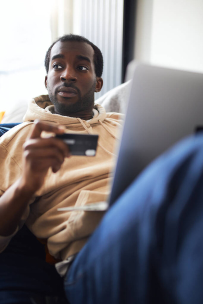 Side view of young mixed ethnicity man lying on sofa at home with laptop computer using credit card to shop online - Фото, зображення