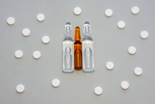 A gyógyszerrel ellátott átlátszó ampullák szürke alapon, fehér kerek tablettákkal körülvéve helyezkednek el. Koncepció fotó illusztrációja kezelés intramuscularis vagy intravénás kábítószer-injekció - Fotó, kép