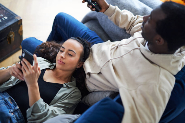 Foto aerea di giovane coppia etnica mista che si rilassa sul divano a casa con l'uomo che guarda la TV mentre la donna si trova in grembo e controlla i social media sul telefono cellulare - Foto, immagini