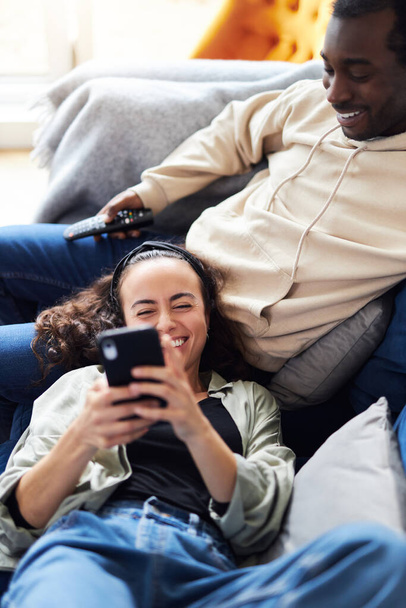 Съемки молодых смешанных этнических пар, отдыхающих дома на диване с мужчиной, смотрящим телевизор, пока женщина лежит на коленях и проверяет социальные сети на мобильном телефоне - Фото, изображение