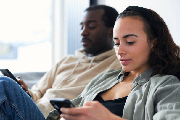 若い混合民族のカップルが自宅でソファに座っている両方の携帯電話でソーシャルメディアをチェックしますが、お互いを無視 - 写真・画像