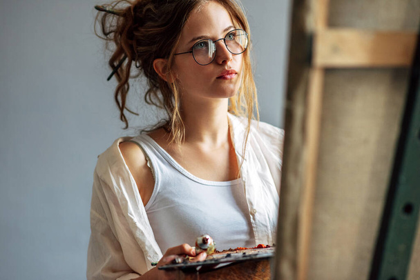Horizontal shot of a girl artist standing next to the easel with canvas painting something in her art studio. A professional young woman painter in transparent eyeglasses draws in the workshop. - Foto, Bild