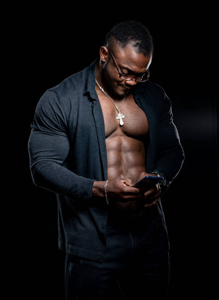 Portrait of a handsome afro american fitness model man. Bodybuilder with unbuttoned shirt shows strong body and types sms on phone. - Foto, Bild