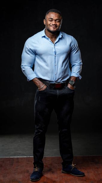 Businessman in dark jeans and light shirt poses to the camera on black background. Full size portrait of an afro american manager. - Foto, imagen