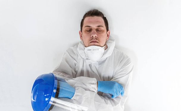 Tired male doctor in a protective suit, sitting on the floor, sleeps exhausted in the fight against a pandemic covid-19, leaning against the wall in a hospital - Zdjęcie, obraz