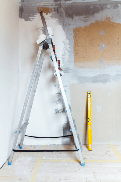 A stepladder with painting tools in a room in a house or apartment. Preparing for putty on the wall or painting. Home repair or renovation concept. - Фото, изображение