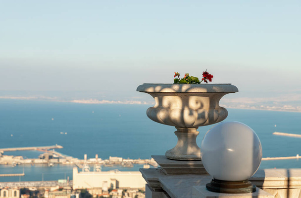 A large stone decorative pot with geraniums and a white lantern on the upper terrace of the Bahai Garden against the backdrop of the downtown and the Mediterranean Sea in the rays of the setting sun in Haifa in northern Israel - Photo, Image