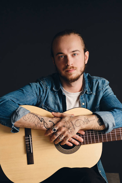 Jovem músico tatuado olhando atentamente na câmera posando com guitarra sobre fundo preto. Elegante cara com guitarra acústica em estúdio - Foto, Imagem