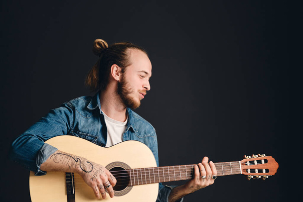 Portrait of tattooed hipster musician playing on guitar and singing over black background. Stylish guy with acoustic guitar in studio - Fotoğraf, Görsel