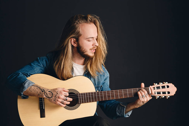 Side view of attractive long haired tattooed guy playing on acoustic guitar and singing preparing for concert isolated on black background. Stylish singer with guitar - Foto, Imagem