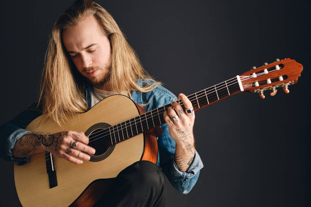 Attraente uomo tatuato dai capelli lunghi che suona su chitarra acustica componendo una canzone isolata su sfondo nero. Elegante cantante maschile con chitarra - Foto, immagini