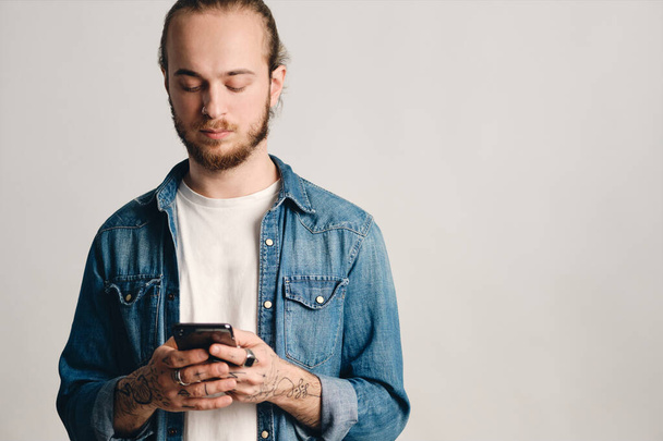 Atraktivní potetovaný chlap oblečený v džínové košili smskování s přáteli pomocí smartphonu izolované na bílém pozadí. Koncept moderní technologie - Fotografie, Obrázek