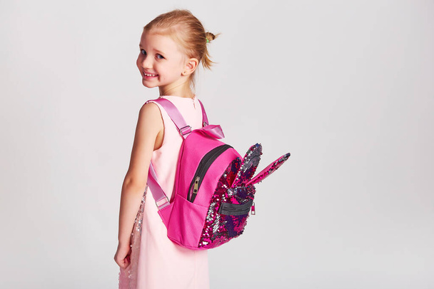 Linda niña con orejas de conejito mochila en vestido de lentejuelas rosa. Concepto de la infancia, la educación y la gente - niña feliz con el bolso de la escuela sobre fondo gris - Foto, Imagen