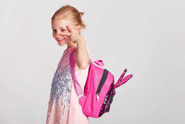 Χαριτωμένο κοριτσάκι με αυτιά λαγού σακίδιο πλάτης σε ροζ φόρεμα με πούλιες. Παιδική ηλικία, εκπαίδευση και άνθρωποι έννοια - χαρούμενος κορίτσι παιδί με σχολική τσάντα πάνω από γκρι φόντο - Φωτογραφία, εικόνα