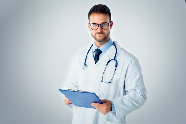 Studio portrait shot of young male doctor holding clipboard in his hand while standing at isolated blue background.  - Foto, Imagen