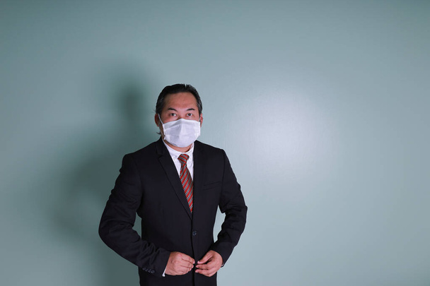 ポートレートアジアのビジネスマンは、医療用マスクを身に着けているカメラを見て黒のスーツを身に着けているコピースペースとコロナウイルスを防ぐことです. - 写真・画像