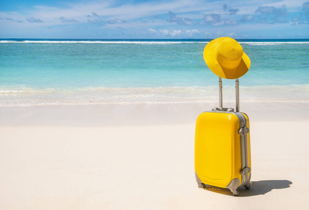 Żółta walizka i kapelusz na pustej tropikalnej plaży z turkusową wodą na tle, letnia tropikalna koncepcja podróży  - Zdjęcie, obraz