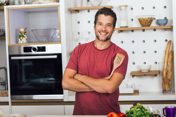 Schöner junger Mann in Freizeitkleidung, der in die Kamera schaut und lächelt, während er zu Hause in der Küche steht - Foto, Bild