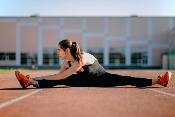 The athletic woman sat down on the splits to warm up before the evening training session at the city stadium. Runner training outdoors, the concept of a healthy lifestyle - Фото, зображення