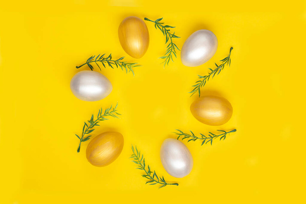 Arany és ezüst tojások, zöld gallyak egy kör alakban, sima sárga háttérrel. Húsvéti üdvözlőlap. Másolótér. - Fotó, kép