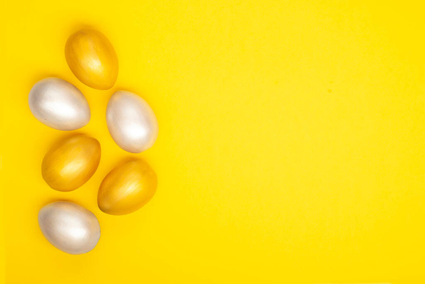 Eier in silberner und goldener Farbe vom linken Bildrand auf einem schlichten gelben Hintergrund. Osterferienkarte. Kopierraum. - Foto, Bild