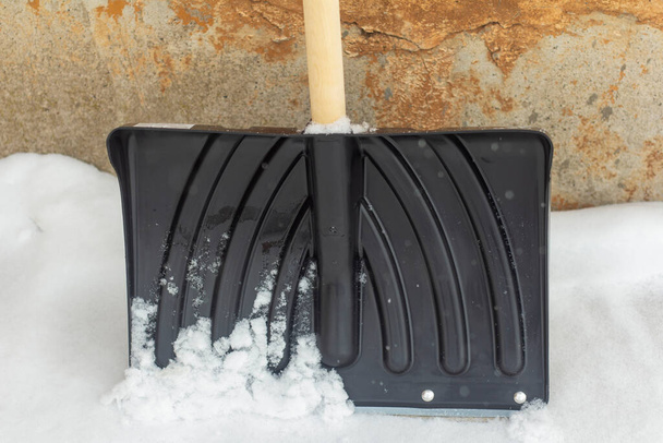 Черная лопата с деревянной ручкой для уборки снега стоит у старой стены. - Фото, изображение