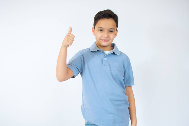 Счастливый ребенок, маленький мальчик показывает большие пальцы вверх жест в голубой футболке изолированы на белом фоне. - Фото, изображение