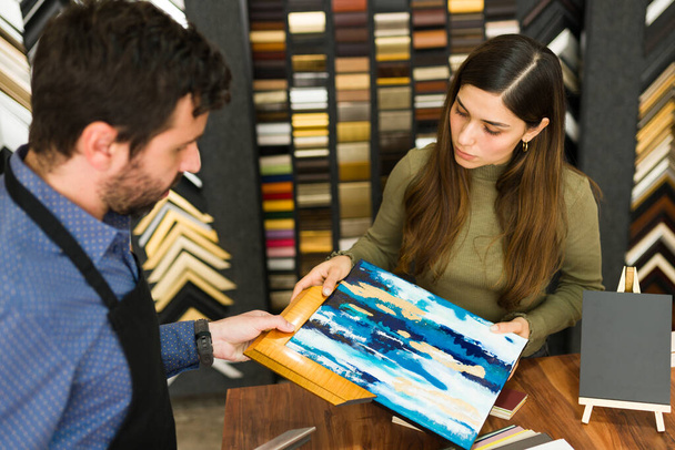 Vendeur caucasien d'un magasin de cadre avec un tablier montre un cadre en bois pour une peinture à un beau client hispanique  - Photo, image