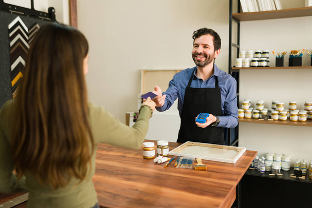 Молода жінка-клієнт передає усміхненому продавцю свою кредитну картку, щоб оплатити свої художні товари в магазині
  - Фото, зображення