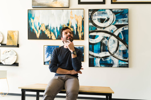 Hombre guapo en sus 30 años con una expresión reflexiva y reflexiva mientras observa las pinturas de arte de una exposición en la galería - Foto, imagen