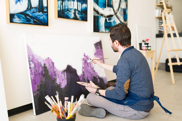 Homme caucasien dans la trentaine vu de derrière assis sur le sol de l'atelier d'art et peignant avec un pinceau une toile colorée  - Photo, image
