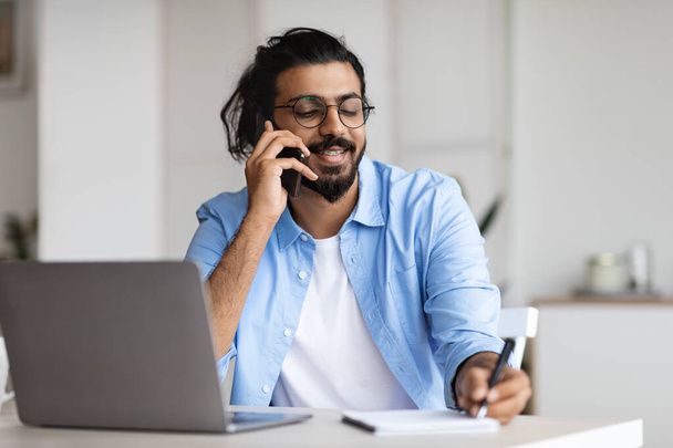 Junge arabische männliche Unternehmerin spricht auf Handy und macht sich Notizen im Büro - Foto, Bild