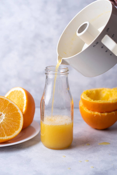 Portakal suyu sıkılmış portakallarla dolu bir masanın üzerindeki meyve sıkacağı şişeye dökülür, hafif bir arka planda, kapatın. - Fotoğraf, Görsel