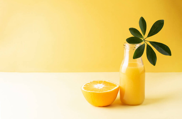 Ένα μπουκάλι φρέσκο χυμό πορτοκαλιού με ένα πράσινο φύλλο και μισό πορτοκάλι σε κίτρινο φόντο, κοντινό πλάνο, αντιγραφή χώρου. - Φωτογραφία, εικόνα