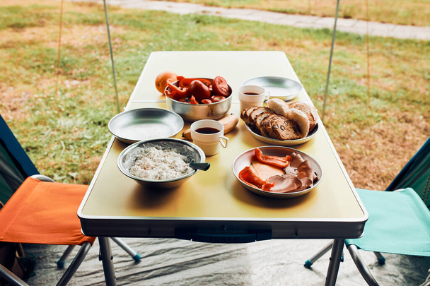 Breakfast prepared during summer vacation on camping. Bread, cottage cheese, cold meat, tomatoes, fruits and coffee cups on table. Close up of outdoor table setting set under tent - Foto, afbeelding