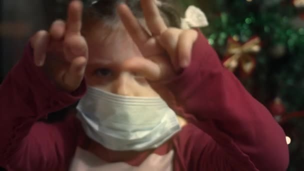 Linda 5 anos. menina mostrando emoções irritadas no fundo da árvore de Natal, descontente por ter que usar uma máscara protetora. Vídeo 4K - Filmagem, Vídeo