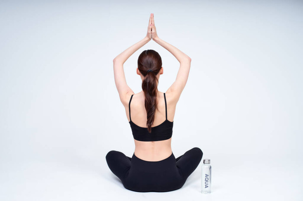 A young healthy girl, in a black top and leggings, practicing yoga, sits in a yoga pose, turning her back, a bottle of water stands next to her. Yoga lady on a white background. Copy space. - Фото, изображение